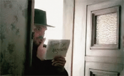 A Man In A Hat Is Standing In Front Of A Door Holding A Piece Of Paper That Says Do N't Say Anything GIF