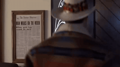 A Man In A Hat Is Standing In Front Of A Framed Newspaper That Says Man Walks On The Moon GIF