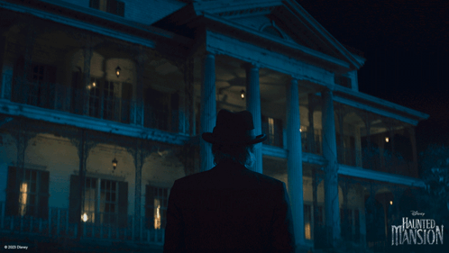 A Man In A Hat Is Standing In Front Of A Haunted Mansion GIF