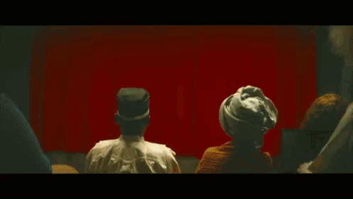 A Man In A Hat Is Standing In Front Of A Red Curtain Looking Out A Window . GIF