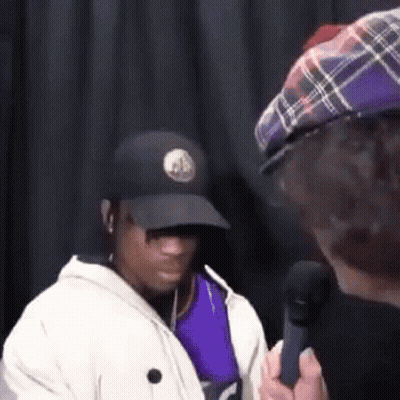 A Man In A Hat Is Talking Into A Microphone While Another Man Looks On . GIF