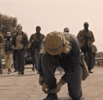 A Man In A Hat Is Tying His Shoelaces While A Group Of People Walk Behind Him GIF