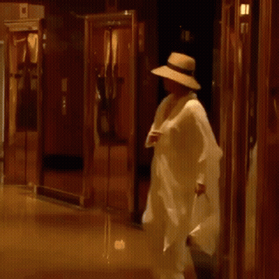 A Man In A Hat Is Walking Down A Hallway Holding A Bag . GIF