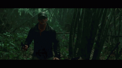 A Man In A Hat Is Walking Through A Dark Forest Holding A Gun GIF
