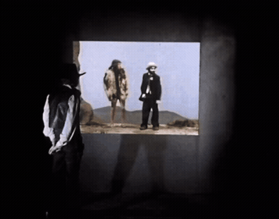 A Man In A Hat Looks Out A Window At Two People GIF