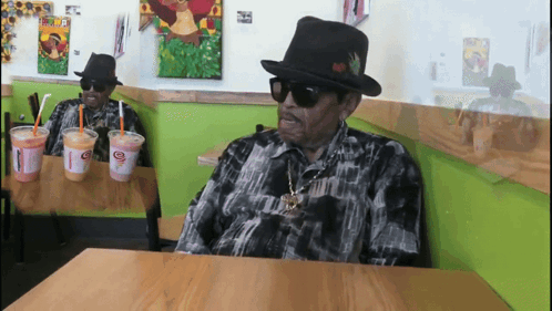 A Man In A Hat Sits At A Table With Two Cups Of Juice GIF