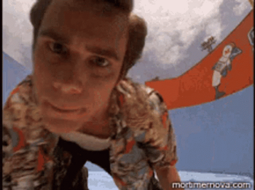 A Man In A Hawaiian Shirt Is Looking Up At The Camera With A Banner In The Background That Says Mortmernova.com GIF