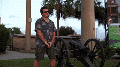 A Man In A Hawaiian Shirt Is Standing Next To A Cannon In A Park GIF