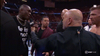 A Man In A He Is Not Shirt Is Talking Into A Microphone While Another Man Looks On . GIF