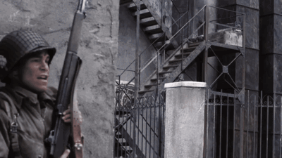 A Man In A Helmet Holds A Gun In Front Of A Building GIF