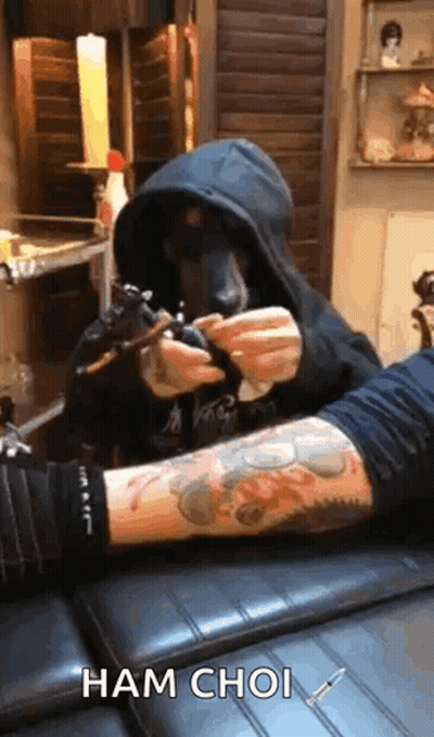 A Man In A Hood Is Getting A Tattoo On Another Man 'S Arm . GIF