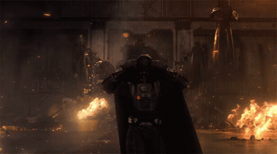 A Man In A Hooded Cape With A Red Light On The Back Stands In Front Of A Fire GIF