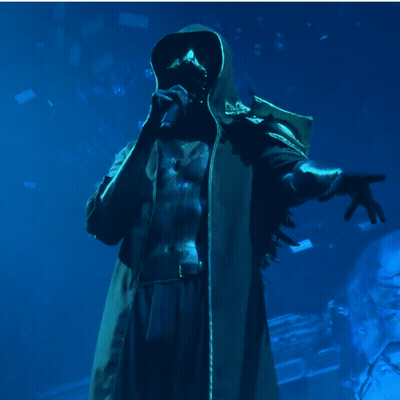A Man In A Hooded Coat Is Singing Into A Microphone In A Dark Room GIF