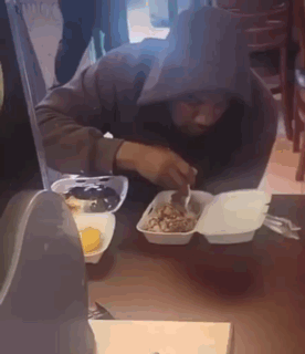 A Man In A Hooded Jacket Is Eating Food From A Styrofoam Container . GIF