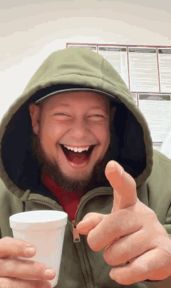 A Man In A Hooded Jacket Is Holding A Cup And Giving A Thumbs Up Sign GIF