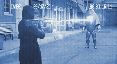 A Man In A Hooded Jacket Is Holding A Gun In Front Of A Camera That Says Cam82 On The Bottom GIF