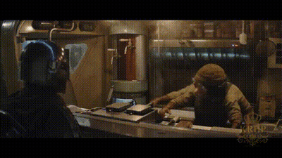 A Man In A Hooded Jacket Is Sitting At A Table In A Dark Room With A Crown On It . GIF