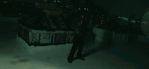 A Man In A Hooded Jacket Stands On A Rooftop Holding An Umbrella And A Microphone GIF