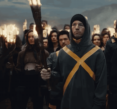A Man In A Hooded Jacket With A Yellow X On It Holds A Torch In Front Of A Crowd GIF