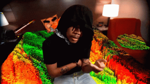 A Man In A Hooded Shirt Is Sitting On A Bed With A Colorful Blanket GIF
