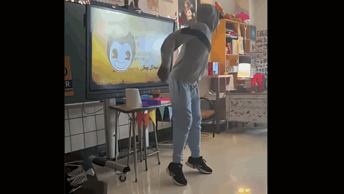 A Man In A Hoodie Is Dancing In A Classroom In Front Of A Large Screen . GIF