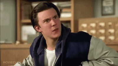 A Man In A Hoodie Is Sitting In Front Of A Bookshelf And Looking At The Camera . GIF