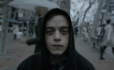 A Man In A Hoodie Is Standing On A Sidewalk With An Umbrella In His Hand . GIF