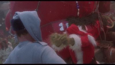 A Man In A Hoodie Talks To A Grinch Who Is Wearing A Santa Hat GIF