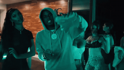 A Man In A Hoodie With A Pendant On His Neck Stands In Front Of Two Women In Bras GIF