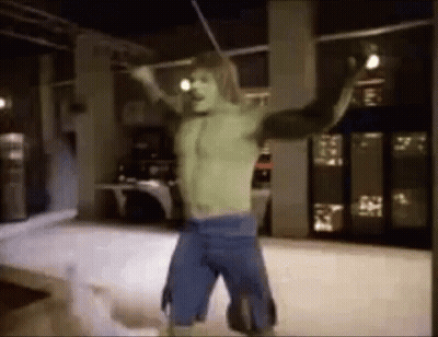 A Man In A Hulk Costume Is Standing In A Room With His Arms Outstretched And Holding A Stick . GIF