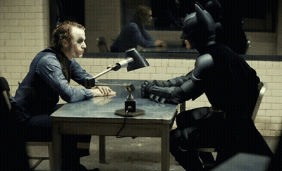A Man In A Joker Costume Sits At A Table With A Batman Costume GIF