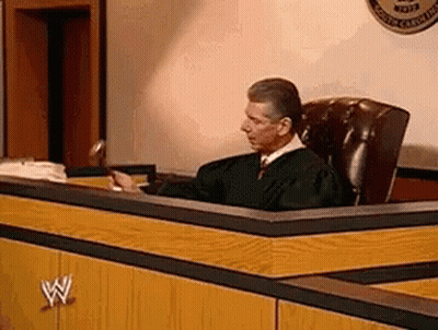 A Man In A Judge 'S Robe Sits At A Desk In A Courtroom . GIF