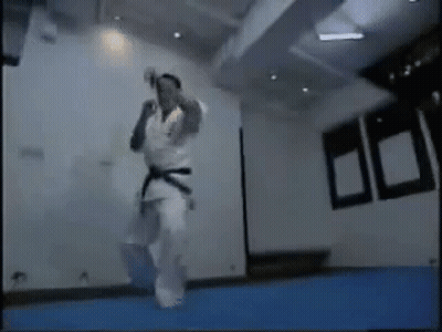 A Man In A Karate Uniform Is Jumping In The Air In A Room . GIF