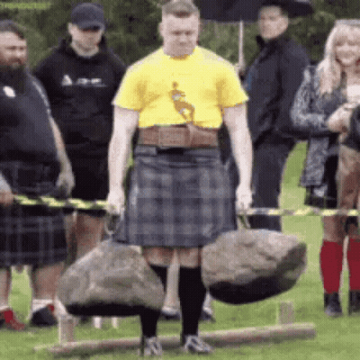 A Man In A Kilt Is Carrying A Large Rock While A Group Of People Watch . GIF