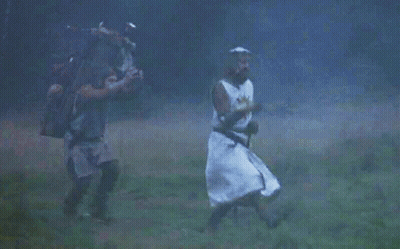 A Man In A Knight 'S Costume Is Walking Through A Field With Another Man Carrying A Backpack . GIF