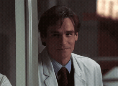 A Man In A Lab Coat And Tie Smiles While Leaning On A Wall GIF