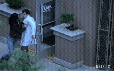 A Man In A Lab Coat Is Being Pushed By A Woman In Front Of A Zalk Building GIF