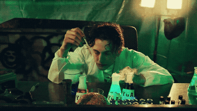 A Man In A Lab Coat Is Sitting At A Table With A Bottle Of Vega On It GIF