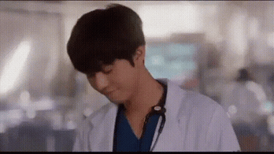 A Man In A Lab Coat With A Stethoscope Around His Neck Is Standing In A Hallway . GIF