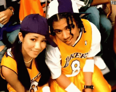 A Man In A Lakers Jersey Sits Next To A Woman Wearing A Purple Hat GIF
