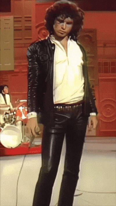 A Man In A Leather Jacket And Pants Stands In Front Of Drums GIF