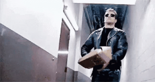 A Man In A Leather Jacket And Sunglasses Is Carrying A Cardboard Box In A Hallway . GIF