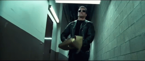A Man In A Leather Jacket And Sunglasses Is Walking Down A Hallway Holding A Piece Of Paper . GIF
