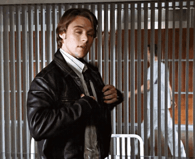 A Man In A Leather Jacket And Tie Is Standing In Front Of A Prison Cell . GIF