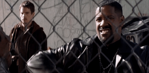 A Man In A Leather Jacket Is Behind A Chain Link Fence With Another Man Behind Him GIF
