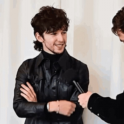 A Man In A Leather Jacket Is Being Interviewed By A Microphone While Standing Next To Another Man . GIF