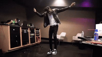 A Man In A Leather Jacket Is Dancing In A Room With A Table And Chairs GIF