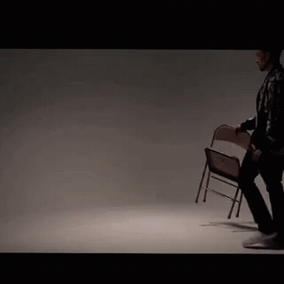 A Man In A Leather Jacket Is Holding A Folding Chair In His Hand . GIF