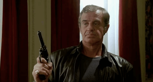 A Man In A Leather Jacket Is Holding A Gun In His Right Hand . GIF