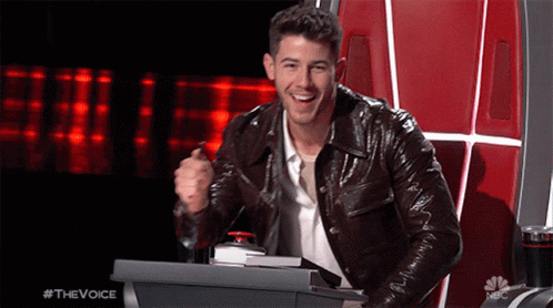 A Man In A Leather Jacket Is Sitting At A Podium With A Red Button That Says #Thevoice GIF